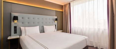A hotel room with a large bed, a nightstand, a wall-mounted lamp, and a window with sheer curtains.
