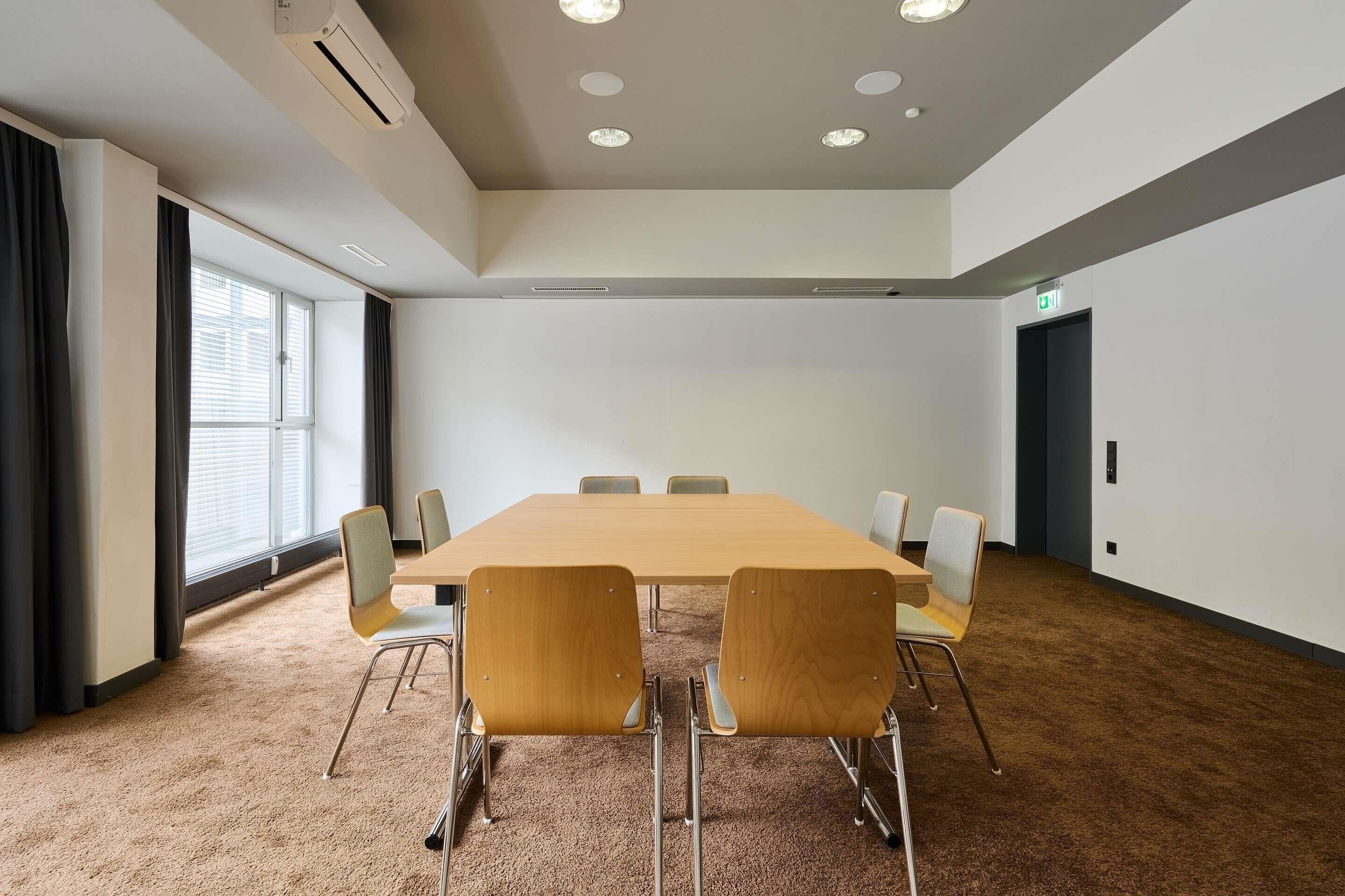 Un salón de conferencias con una mesa larga, seis sillas, suelo alfombrado, luces de techo y una ventana con cortinas.