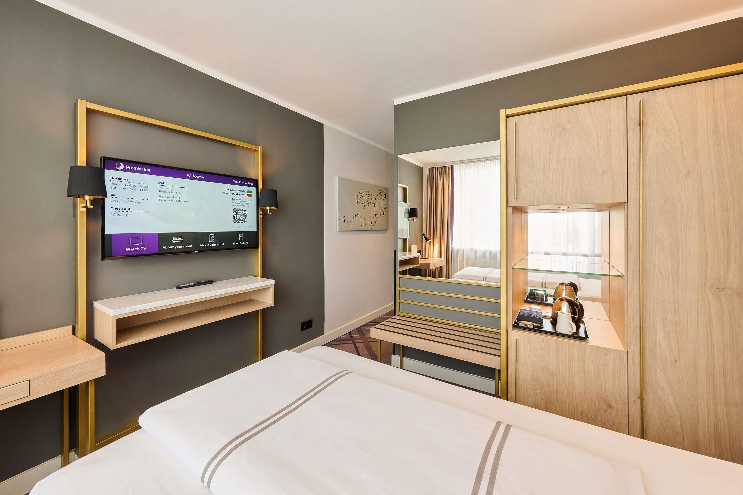 A hotel room with a bed, a TV mounted on the wall, a desk with a lamp, and a wardrobe.