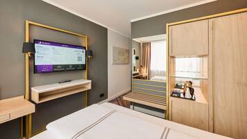 A hotel room with a bed, a TV mounted on the wall, a desk with a lamp, and a wardrobe.