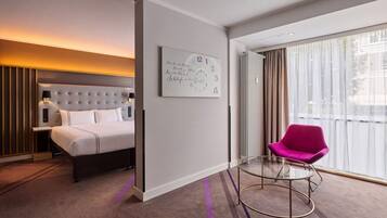 A modern hotel room with a bed, a chair, a bedside table, a wall clock, and a large window with curtains.