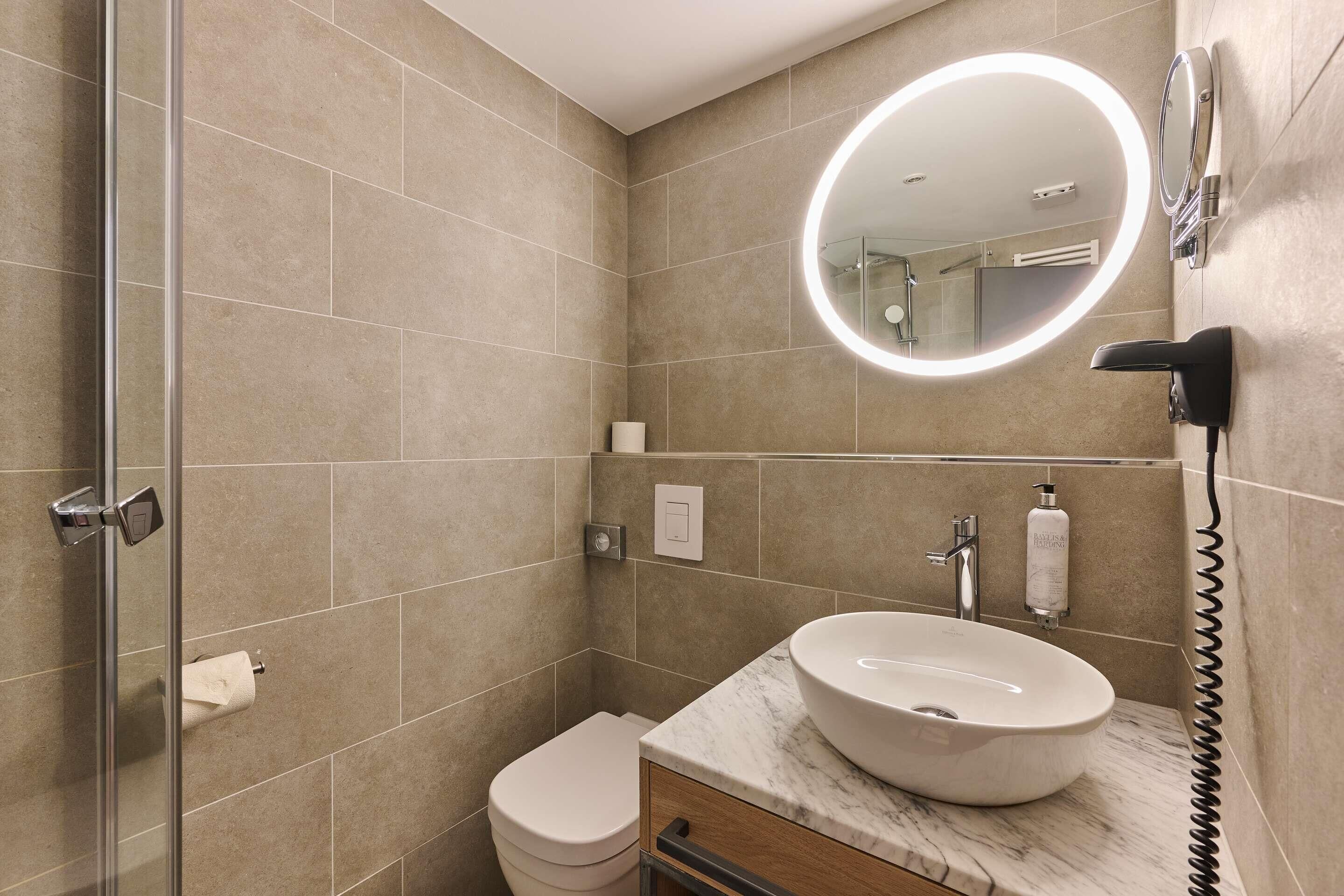 A bathroom with a glass shower enclosure, a toilet, a sink with a marble countertop, and a wall-mounted mirror with a light.