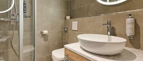 Ein modernes Badezimmer mit Duschkabine aus Glas, einer weißen Toilette, einem großzügigen Waschtisch und einem Spiegel mit runder Beleuchtung.