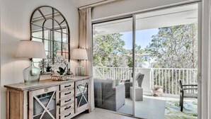 Ein Zimmer mit einem Holzkoffer, zwei Lampen, einem Spiegel und einer Glastür, die auf einen Balkon mit Korbmöbeln und Blick auf Bäume führt.