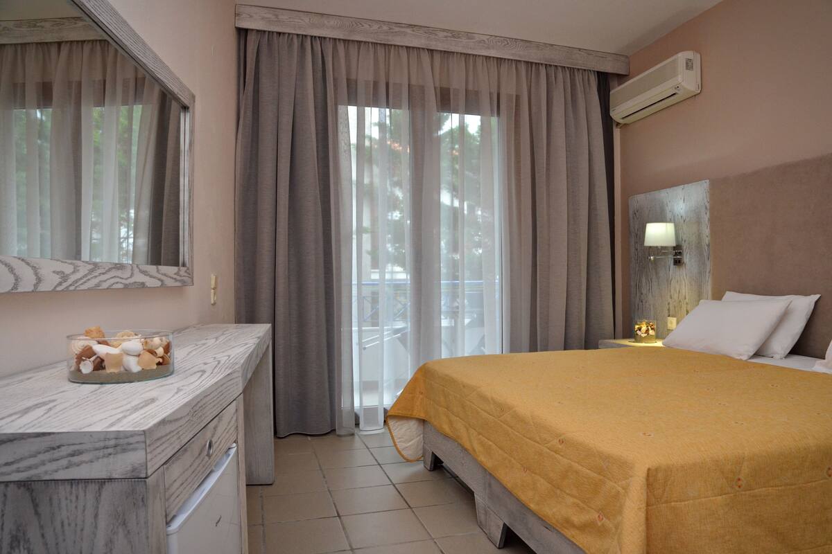 A hotel room with a bed, a marble bedside table, a nightstand with a lamp, and a view through the window.
