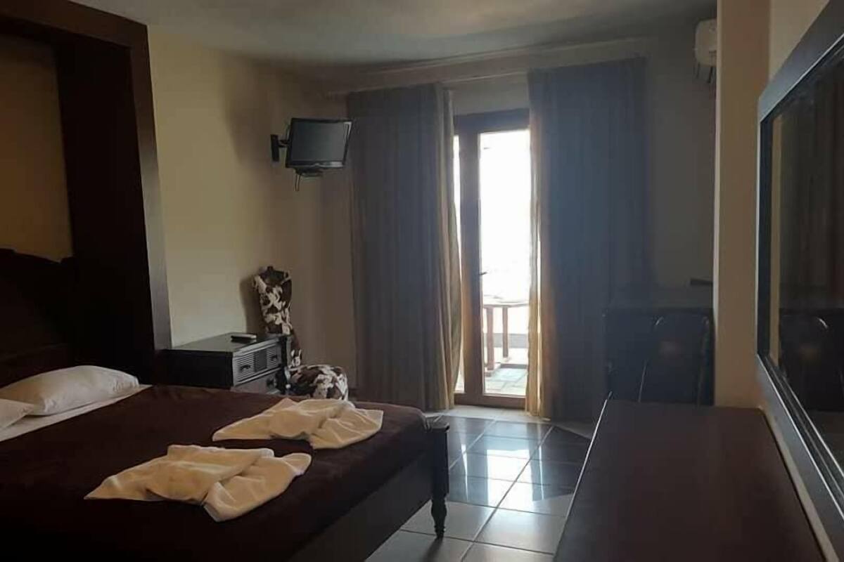 A hotel room with a large bed, a flat-screen TV mounted on the wall, a small table, and a window with curtains.