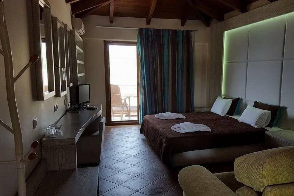 A hotel room with a bed, a desk with a computer, a chair, a bedside table, and a large window with curtains.