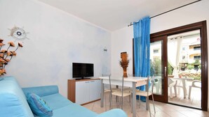A modern living room with a blue sofa, a TV on a cabinet, a dining table with chairs, and a view through a glass door to an outdoor area. - Two-room apartment with air conditioning and swimming pool (Santa Teresa Gallura)