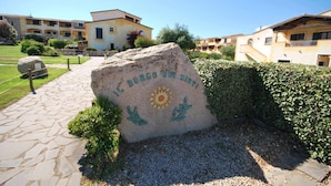 A stone plaque with text and a sun emblem, surrounded by a gravel path and greenery, with buildings in the background. - Two-room apartment with air conditioning and swimming pool (Santa Teresa Gallura)