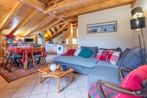 A cozy living room with a sofa, a coffee table, and a dining area with a table and chairs. There is a kitchen in the background with a washing machine and a sink.