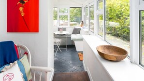 Una habitación luminosa e inundada de sol, con techo y paredes blancos, un ventanal con vistas a zonas verdes, una silla rojo y blanco, y un cuenco de madera en el alféizar.