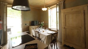 A dining area with a table set for two, a large window with curtains, and a wooden door. - Apartment Alba 4 in Villa Giobatta (Barolo)