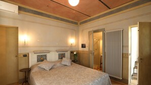 A hotel room with a large bed, a small table, a radiator, and a door leading to a bathroom. - Apartment Alba 4 in Villa Giobatta (Barolo)