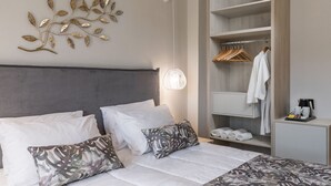 Une chambre d’hôtel comprenant un lit, deux oreillers, une table de chevet avec une machine à café, une armoire avec des vêtements suspendus et un élément décoratif en forme de feuille fixé au mur.