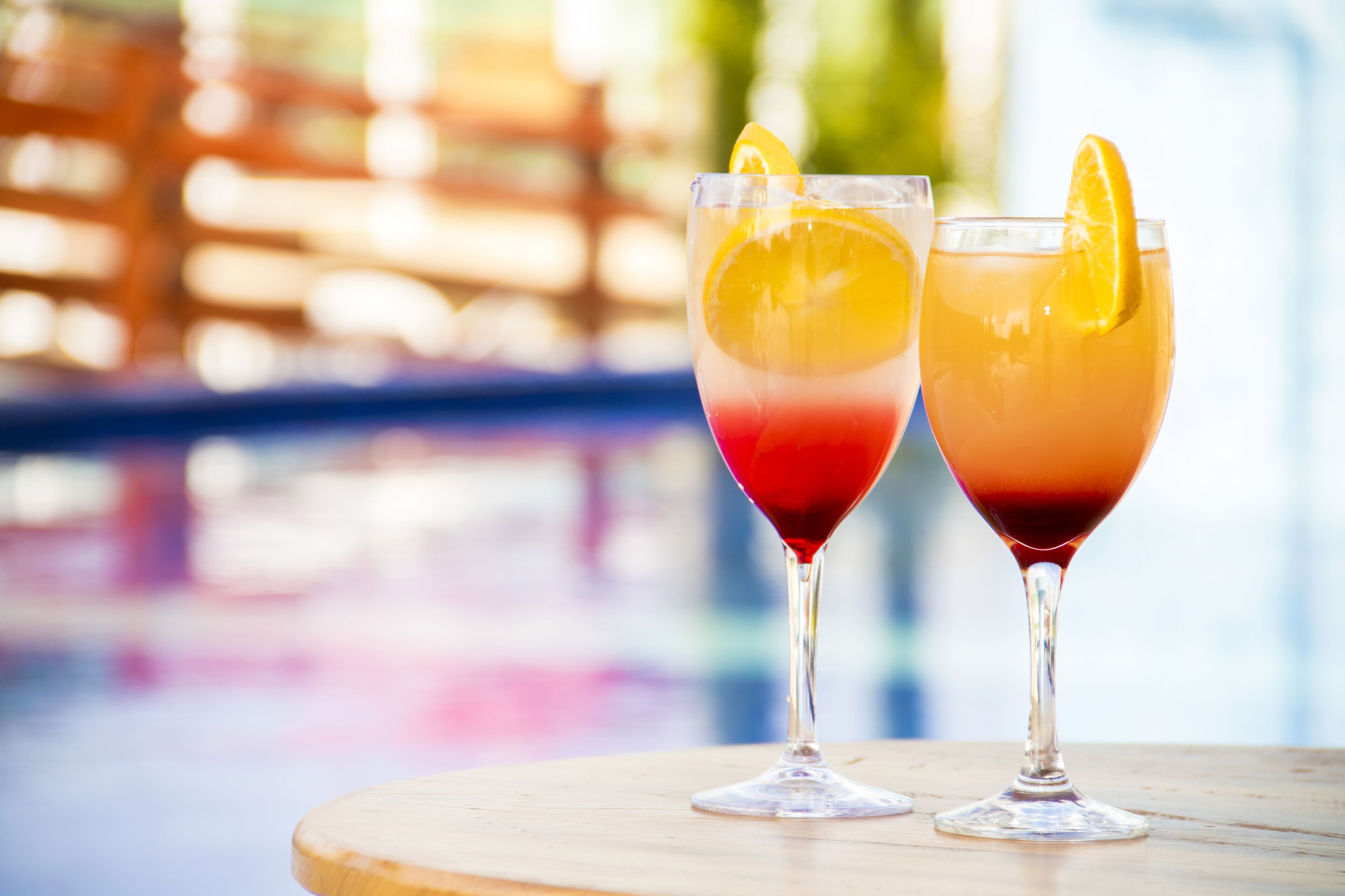 Two cocktails on a wooden table with a blurred background of a pool and a wooden fence.