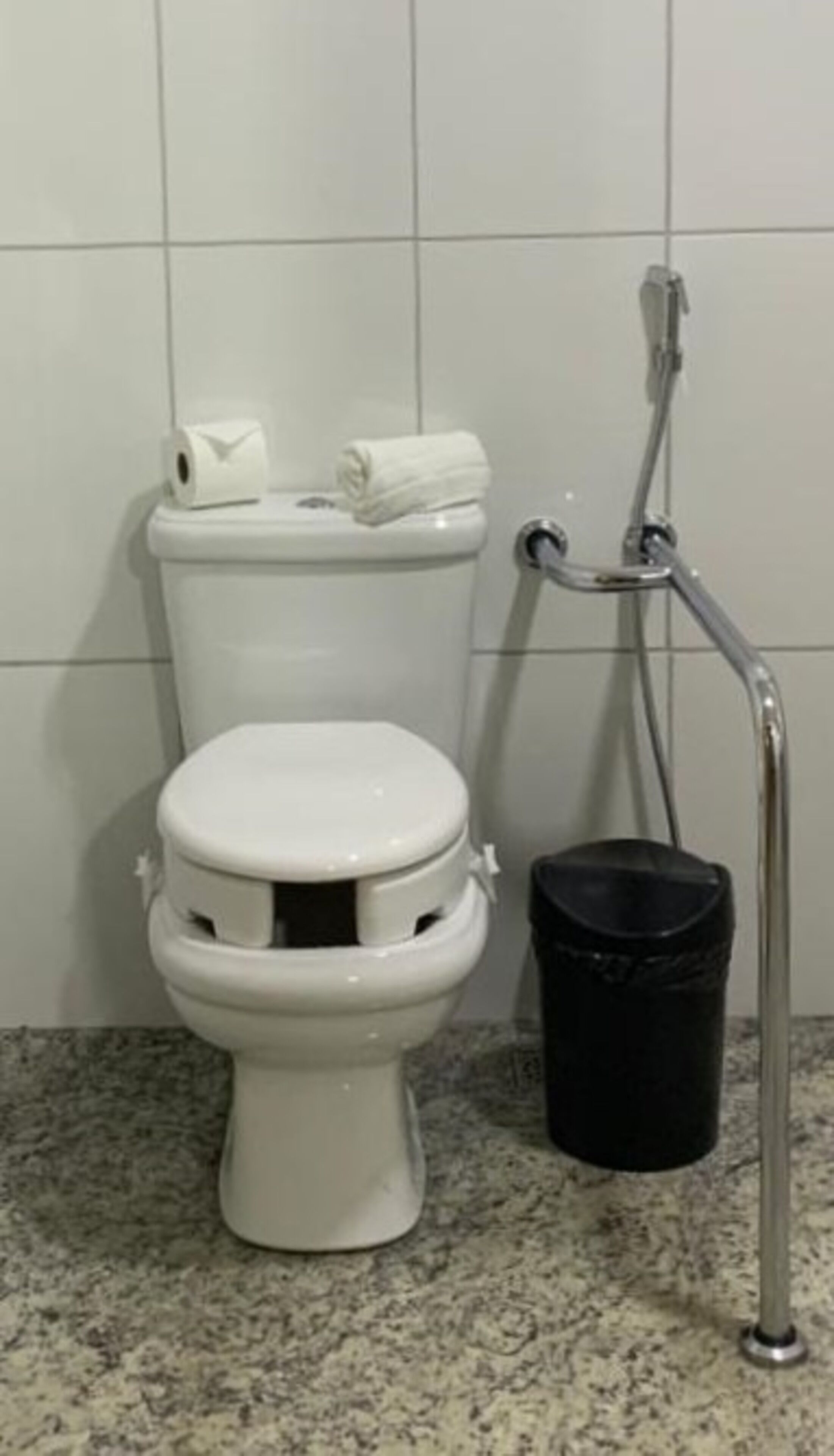 A toilet with a roll of toilet paper and a towel on the tank, a grab bar, and a black trash bin.