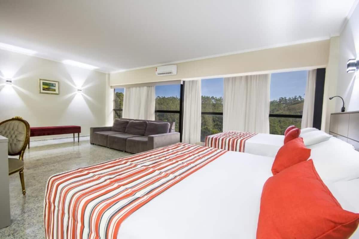 A hotel room with a large bed, a sofa, a chair, and a small table. There are large windows with curtains, and a painting on the wall.