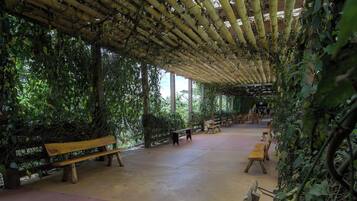 An outdoor covered area with benches and tables, surrounded by dense foliage.
