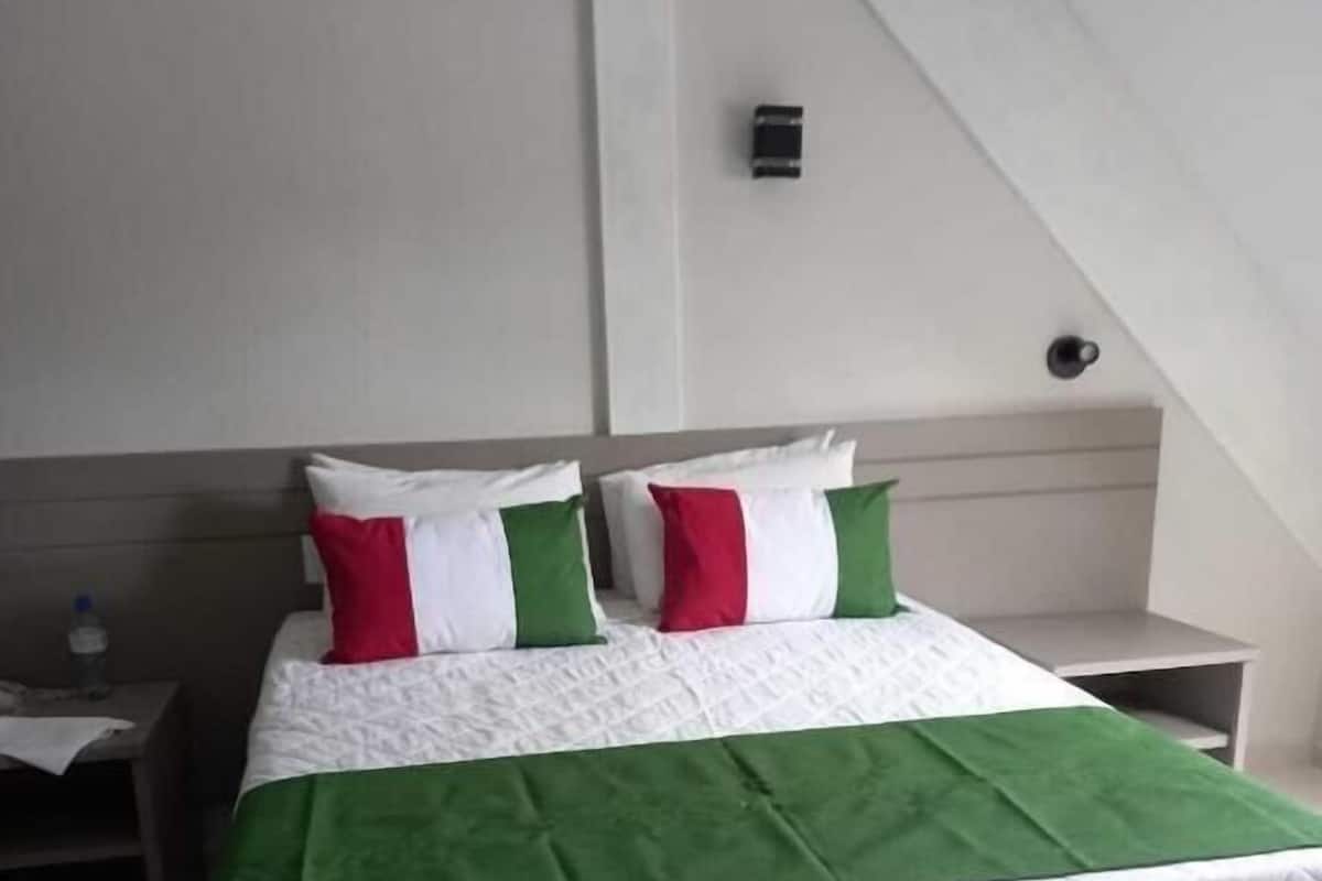 A neatly made bed with a green and white bedspread and pillows, a white nightstand with a water bottle, and a white wall with a small light fixture.