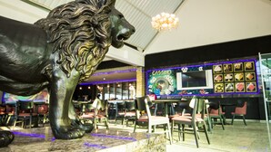A lion statue in a spacious indoor area with tables and chairs, a large screen displaying casino games, and a chandelier.