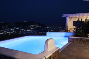 A nighttime view of a swimming pool with a cityscape in the background. - Villa Aella (Mykonos)