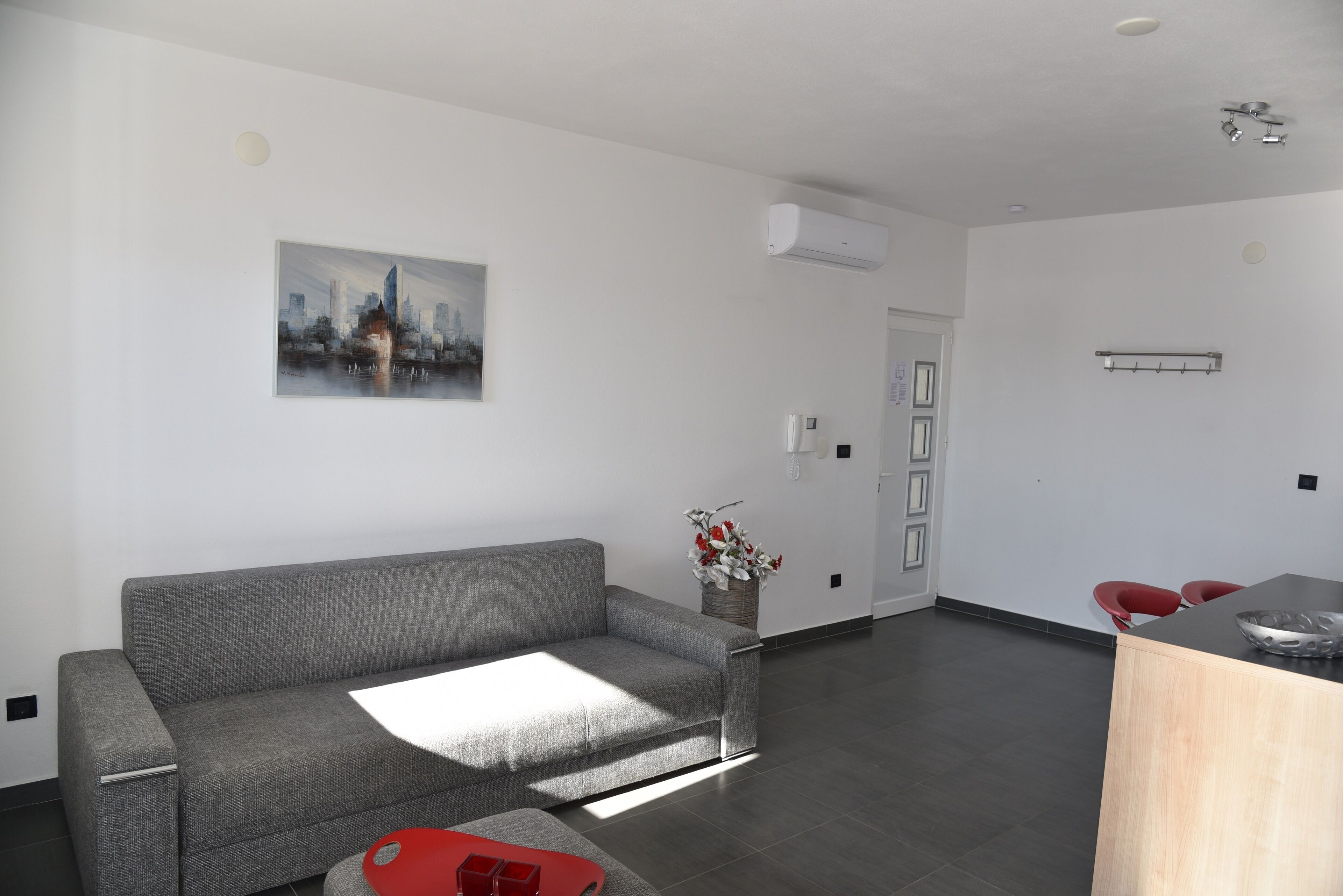 Un salon moderne avec un canapé gris, un fauteuil rouge, une table basse en bois et un tableau représentant un paysage urbain accroché au mur.