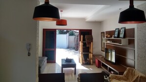 Ein Wohnzimmer mit einer Couch, einem Couchtisch und einem Fernseher auf einem Holzständer. Zwei Pendelleuchten hängen von der Decke.