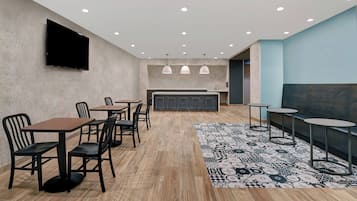 Un espace repas moderne avec des tables et des chaises, un tapis à motifs, des luminaires suspendus et un téléviseur à écran plat fixé au mur.