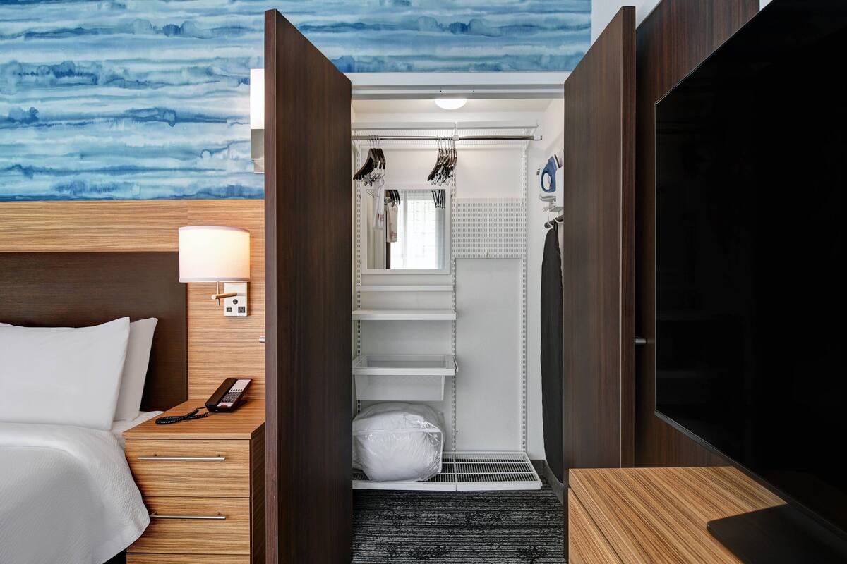 Une chambre d’hôtel comprenant un lit, un bureau avec une lampe, une télévision, une armoire, une salle de bain avec douche et un mur orné d’un motif abstrait bleu.