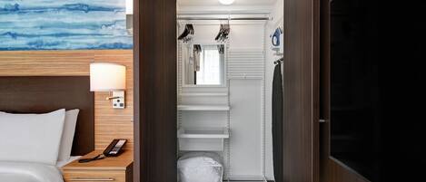 A hotel room with a bed, a desk with a lamp, a TV, a wardrobe, a bathroom with a shower, and a wall with a blue abstract design.
