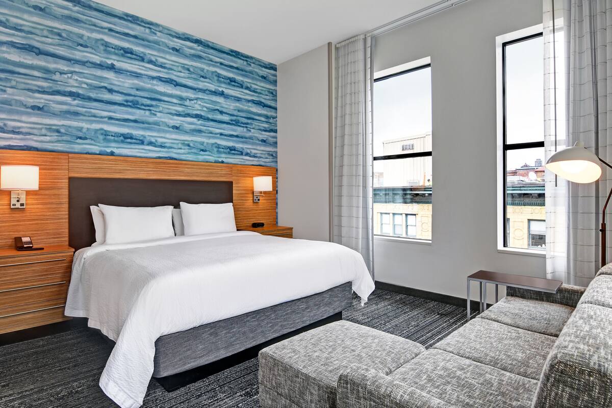 Une chambre d’hôtel avec un grand lit, une tête de lit en bois, deux lampes de chevet, un canapé gris recouvert de tissu et une vue sur la ville à travers de grandes fenêtres.