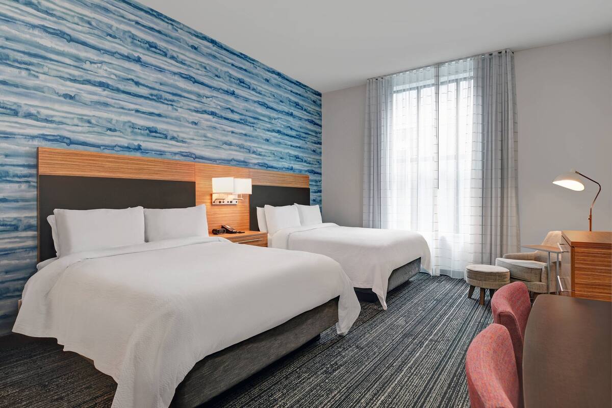 Une chambre d’hôtel avec deux lits, un bureau, une chaise, une lampe et une fenêtre avec des rideaux.