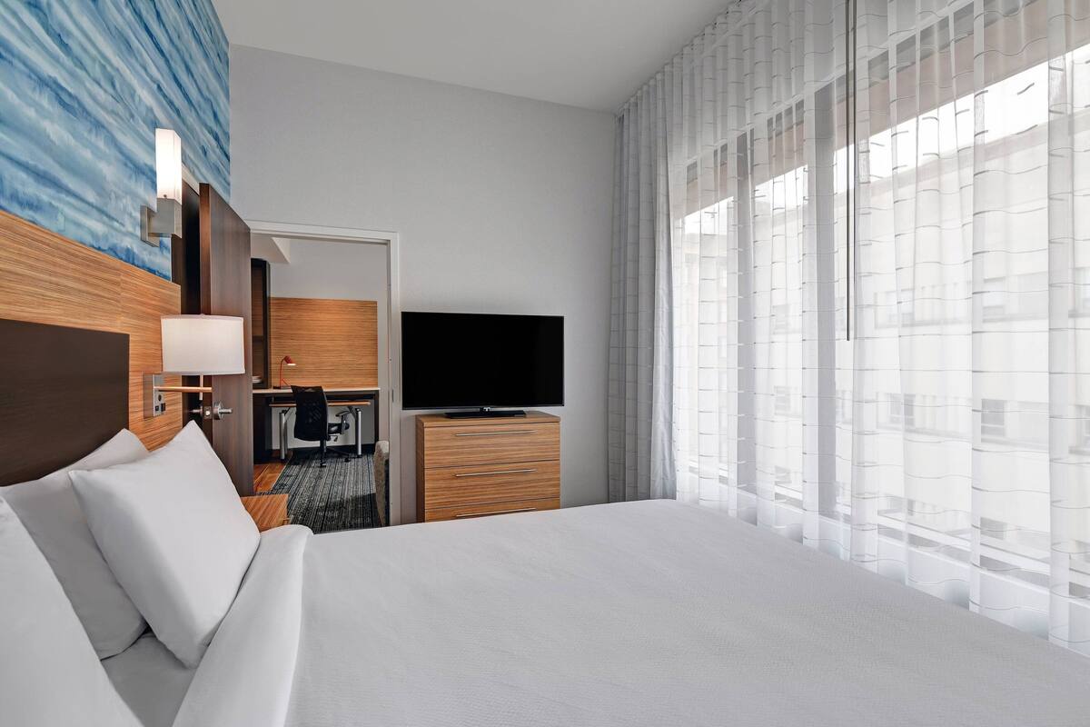 Une chambre d’hôtel avec un grand lit, un téléviseur à écran plat, une commode en bois, un bureau avec une chaise et une vue sur les bâtiments à travers de fines rideaux.