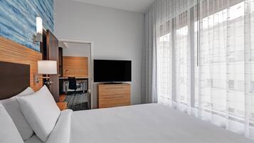 Una camera d'albergo con un letto grande, una televisione a schermo piatto, una cassettiera in legno, una scrivania con una sedia e vista sugli edifici attraverso tende trasparenti.