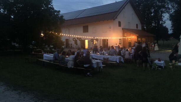 An outdoor event with tables and chairs arranged in front of a building with a sloped roof and string lights.