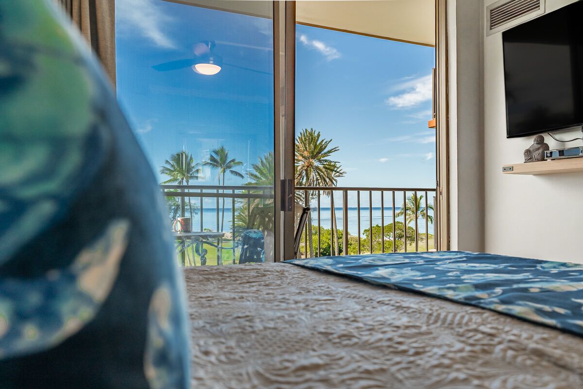 A room with a large sliding glass door leading to a balcony with a view of palm trees and the ocean.