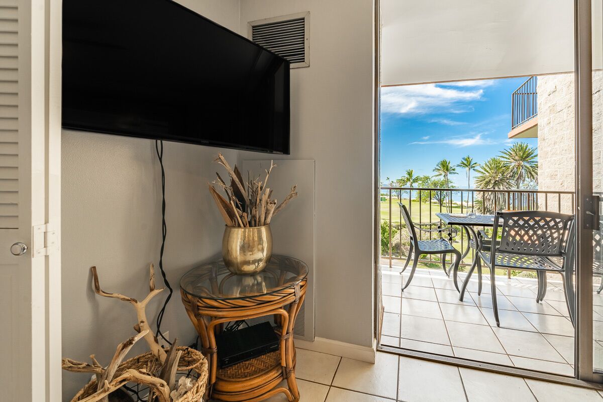 Ein Zimmer mit an der Wand montiertem Fernseher, einem Korbstuhl-Nachttisch mit einer Vasen-Arrangement aus Zweigen und Blick auf eine Außenpatio mit Tisch und Stühlen.
