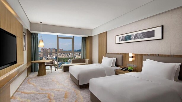 Habitación de hotel con dos camas, televisión, escritorio y vista a la ciudad.