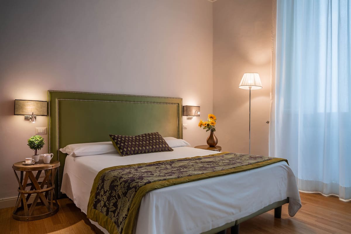 A neatly made bed with a green headboard, a bedside table with a lamp, and a small potted plant.