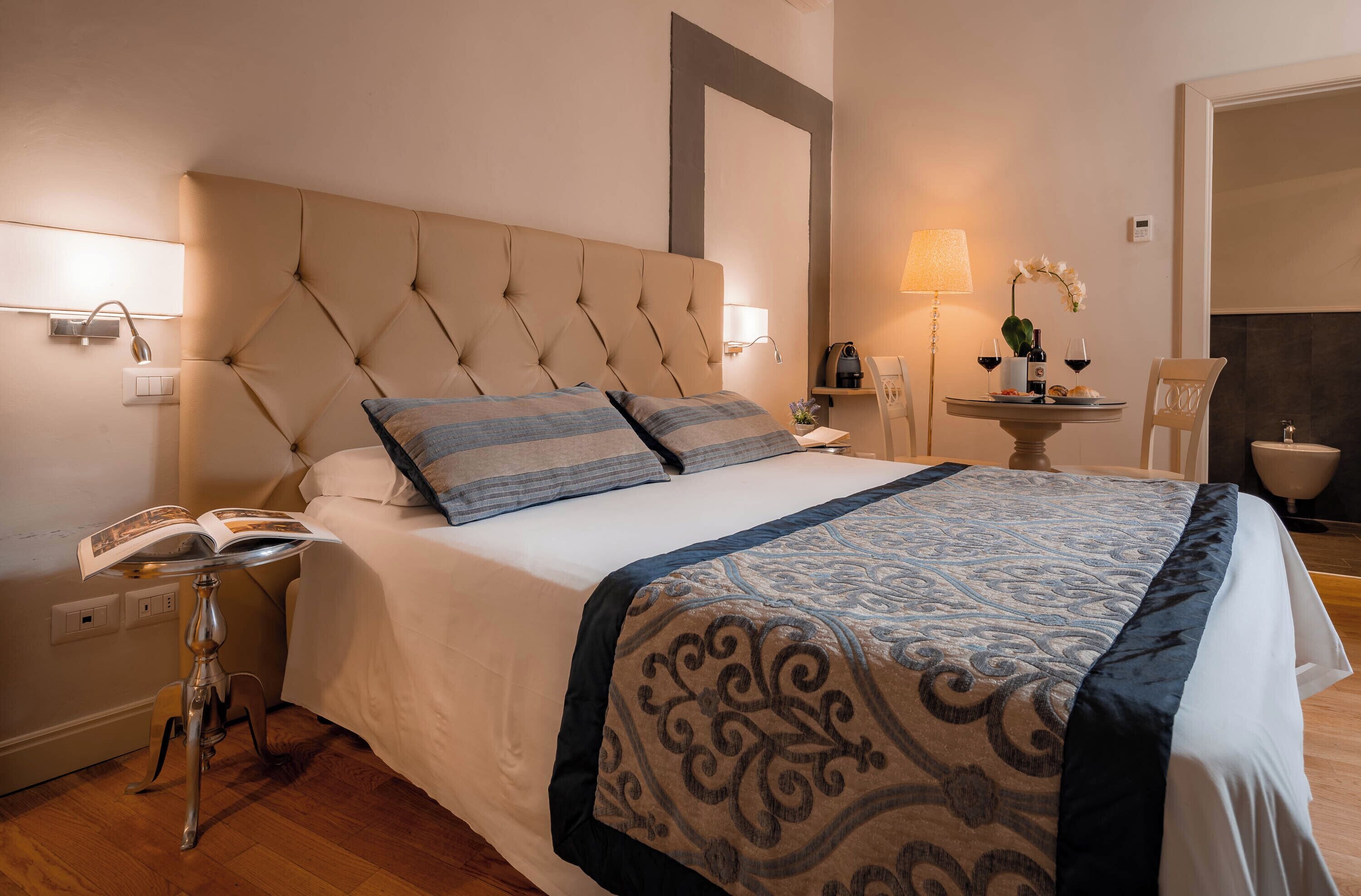 A neatly made bed with a patterned bedspread, a bedside table with a lamp, and a small table with a vase of flowers.