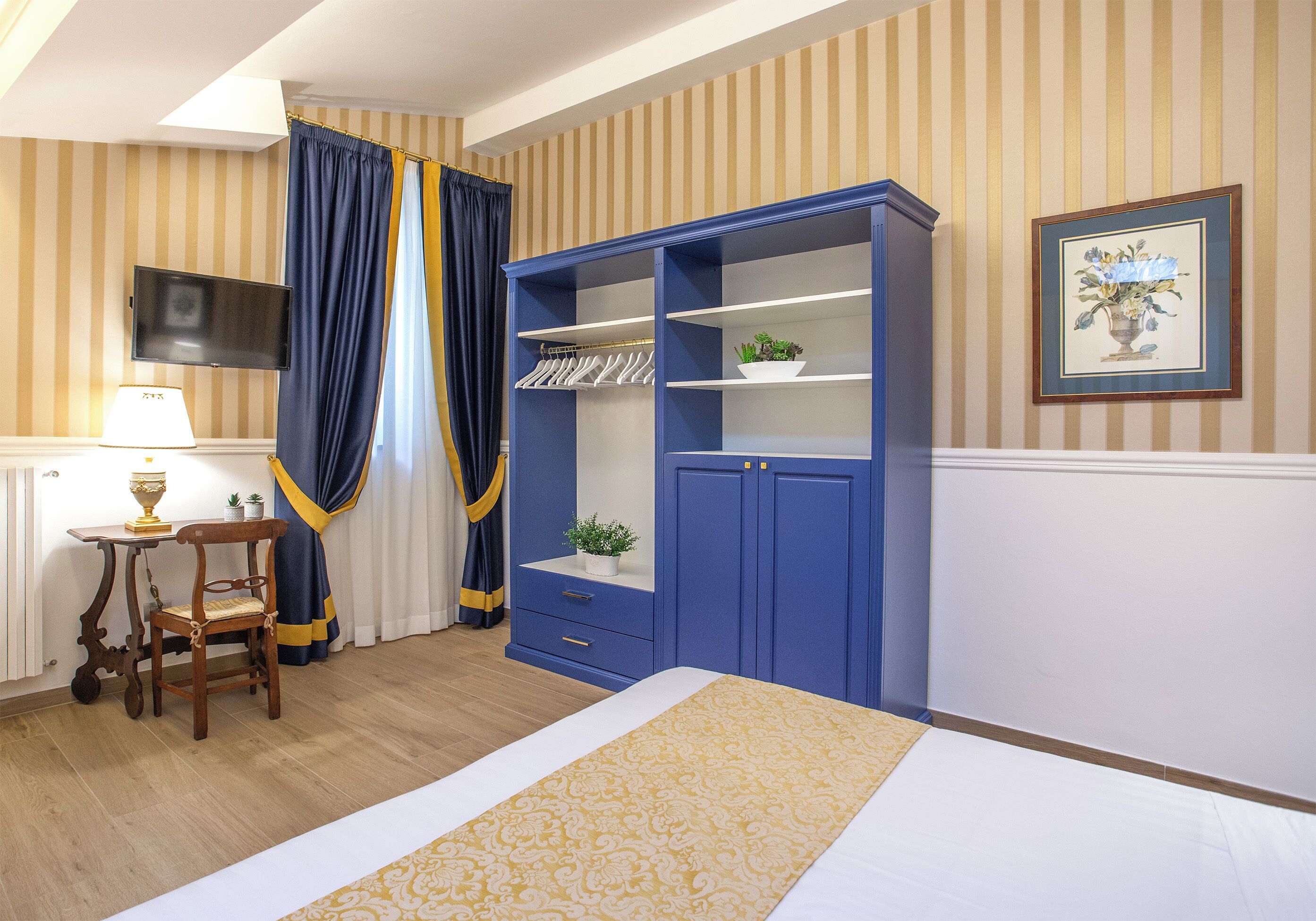 A hotel room with a bed, a wardrobe, a desk with a lamp, a chair, and a television on the wall.