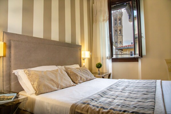 A hotel room with a striped wall, a bed with white linens, a nightstand with a lamp, and a view of a tower through a window. - Boutique Hotel del Corso  (Florence)