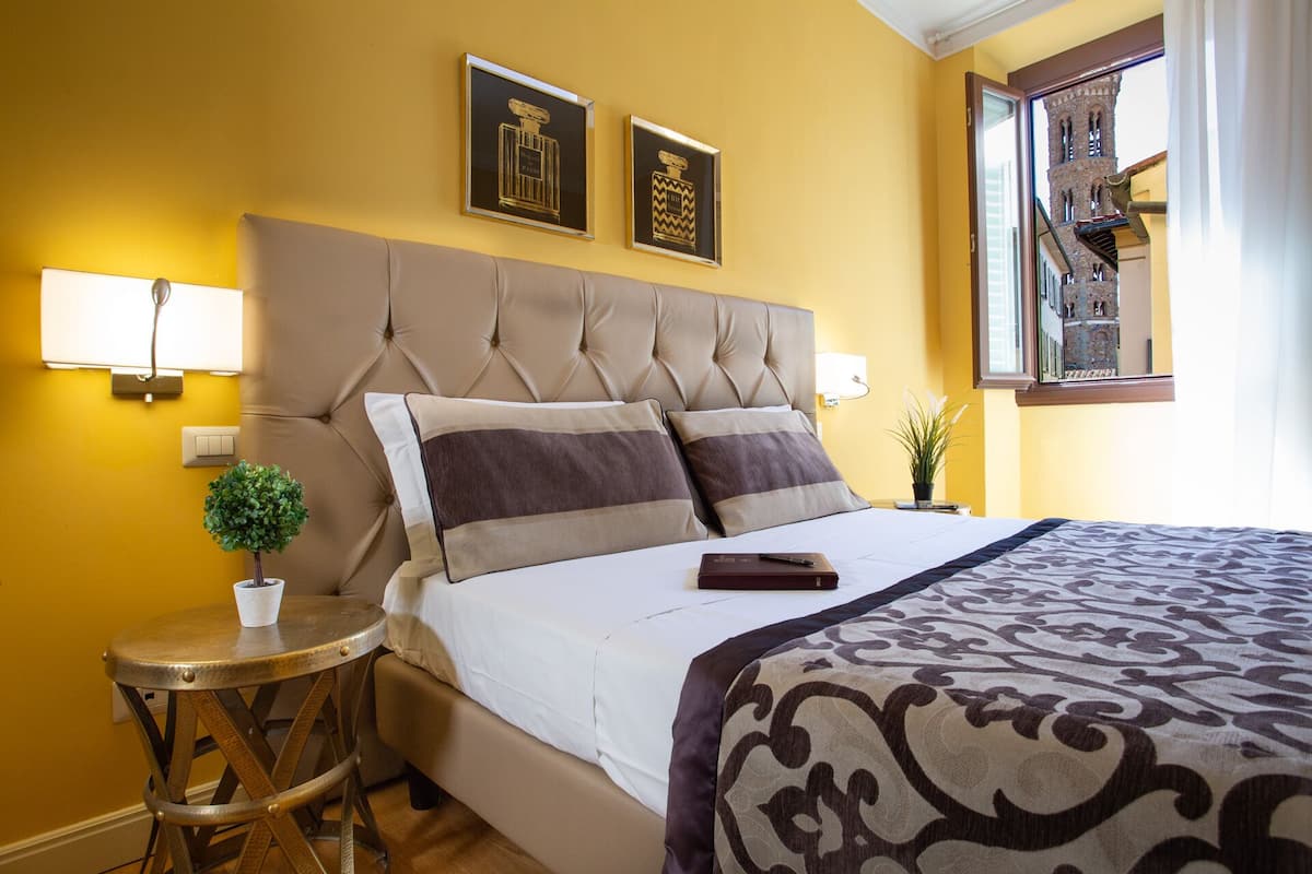 A hotel room with a tufted headboard, two framed perfume bottles, a small round table with a potted plant, and a view of a historic building through a window.