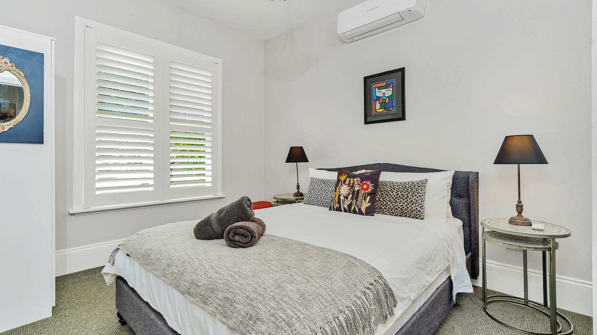 A bedroom with a large bed, two bedside tables with lamps, a window with blinds, and a wall-mounted air conditioner.