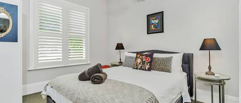 A bedroom with a large bed, two bedside tables with lamps, a window with blinds, and a wall-mounted air conditioner.