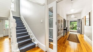A staircase with a dark carpeted finish leading to a white door. A hallway with wooden flooring and a rug is visible, leading to a door with a view of greenery outside.