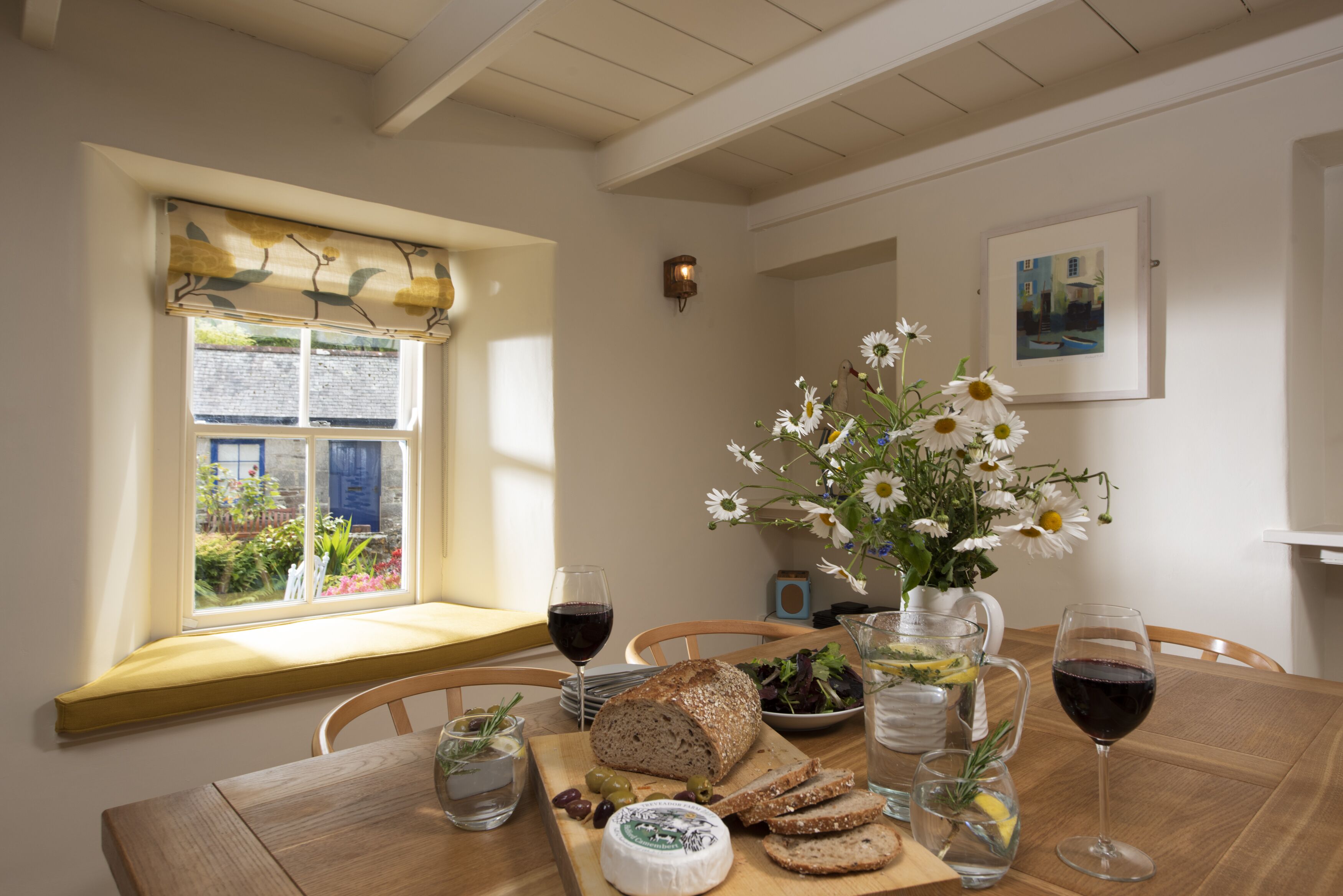 A dining table with a loaf of bread, cheese, olives, and a vase of flowers.