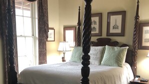 Een hemelbed met een witte spreidek en een aansluitende onderrok. Er zijn twee nachtkastjes, waarvan er eentje een lamp heeft en de ander een telefoon. De kamer heeft een raam met gordijnen en een ingelijste foto aan de muur.