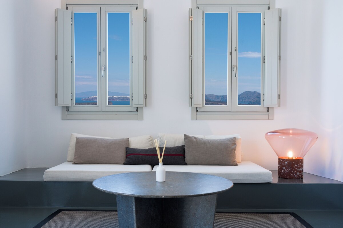 A modern room with a sofa, a round table, and two windows with a view.