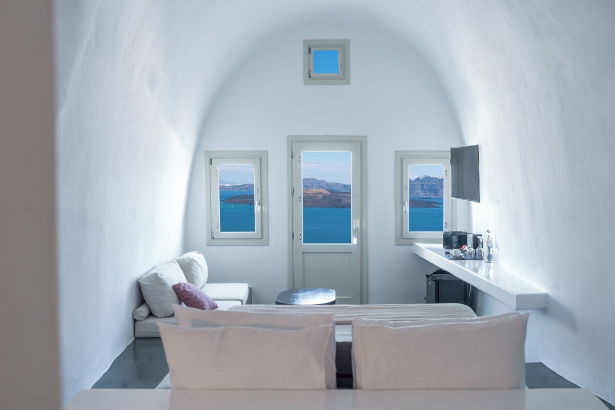 Une pièce dotée d’un plafond voûté, d’un lit, d’un canapé, d’une petite table et de trois fenêtres offrant une vue sur la mer et les montagnes.